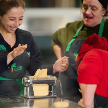 Two women laughing together while making fresh pasta on a hand-cranked pasta machine at a community cooking class